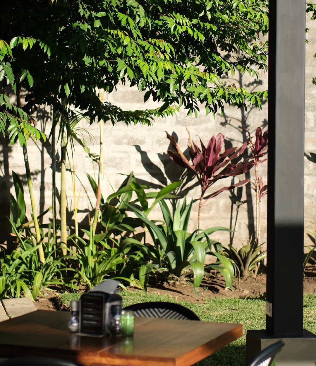 El jardín de Wö Kàpi con bambú, plantas tropicales y una mesa de madera a la sombra de un árbol