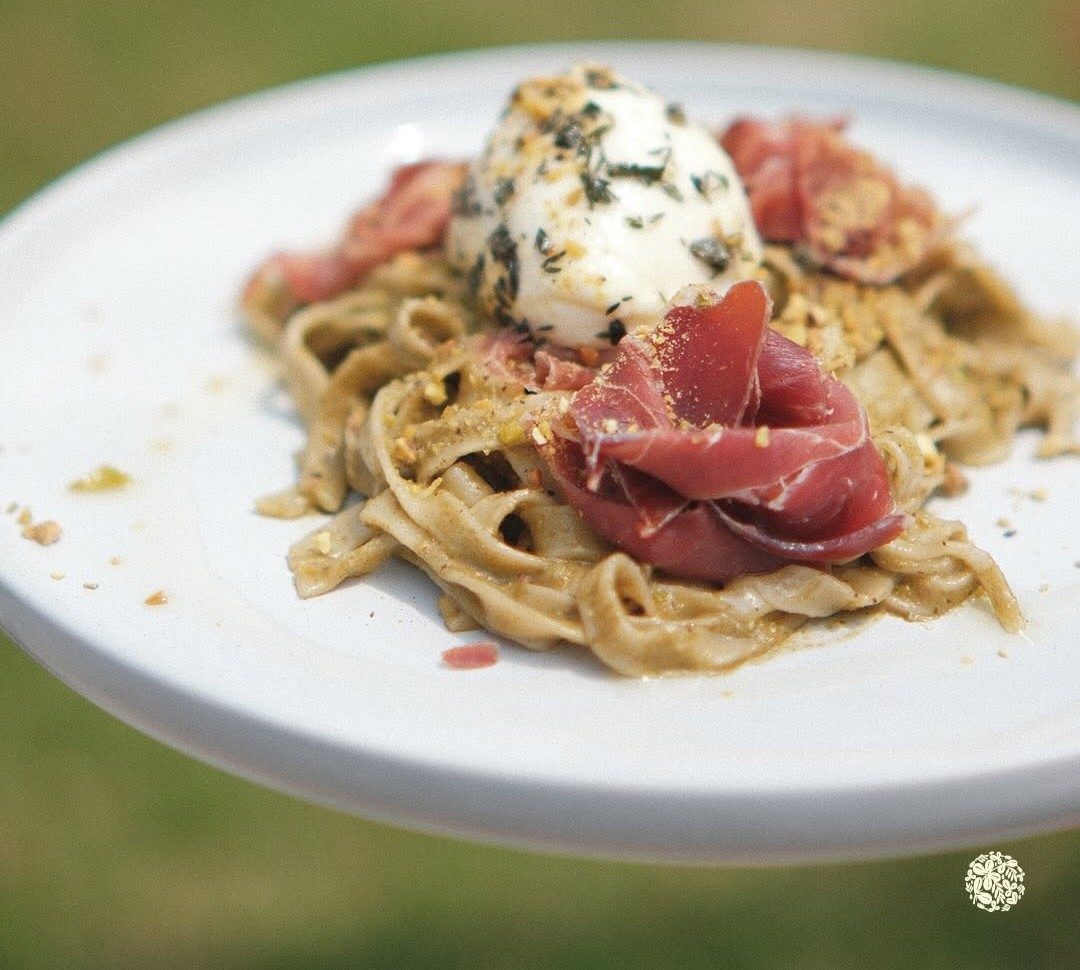 Pasta con pesto de pistacho, jamón serrano y burrata sostenida en alto sobre el verde del jardín
